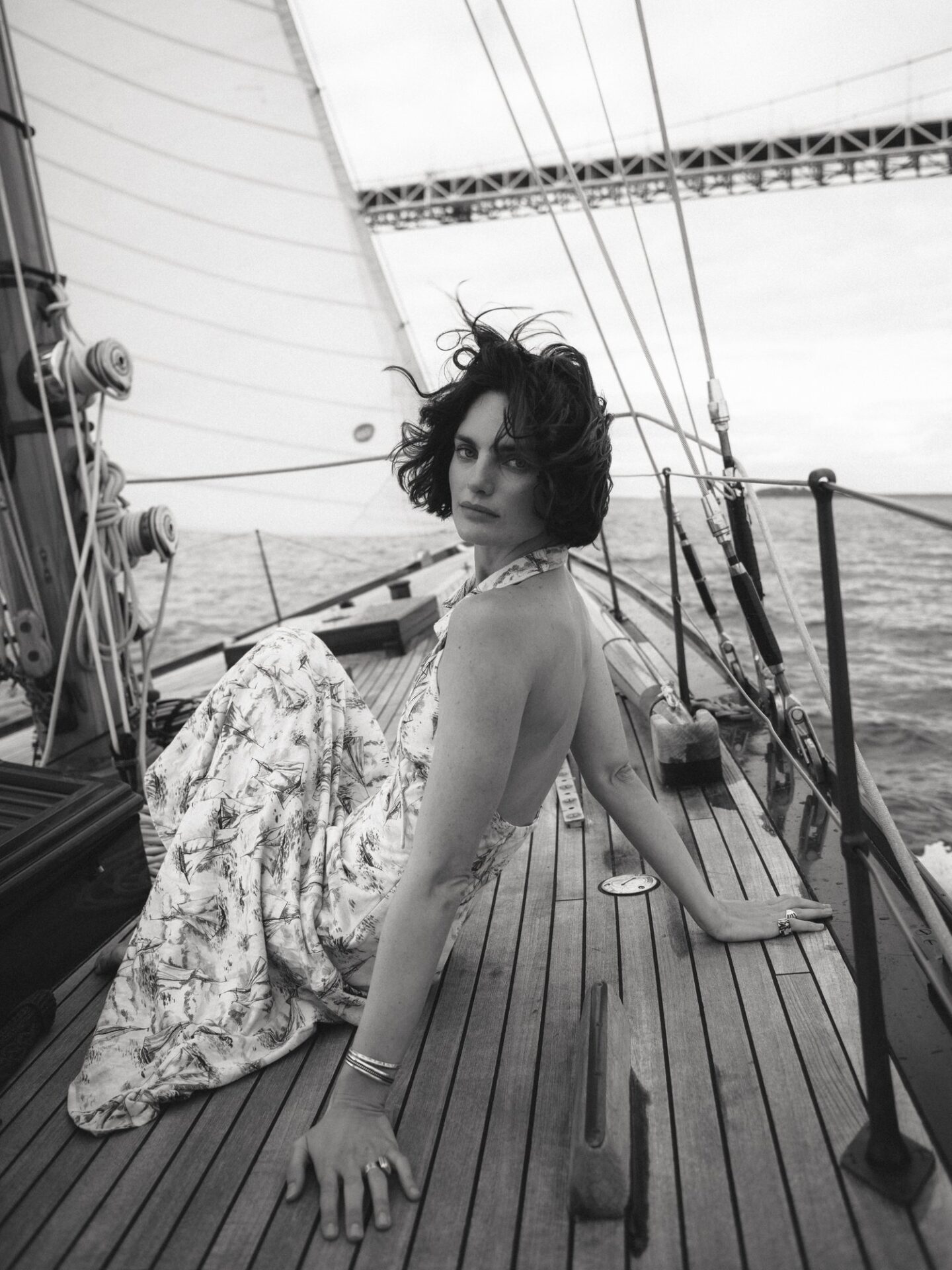 Black and white shot of a woman in a patterned dress sitting on a wooden sailboat deck, with sail and water visible in the background.