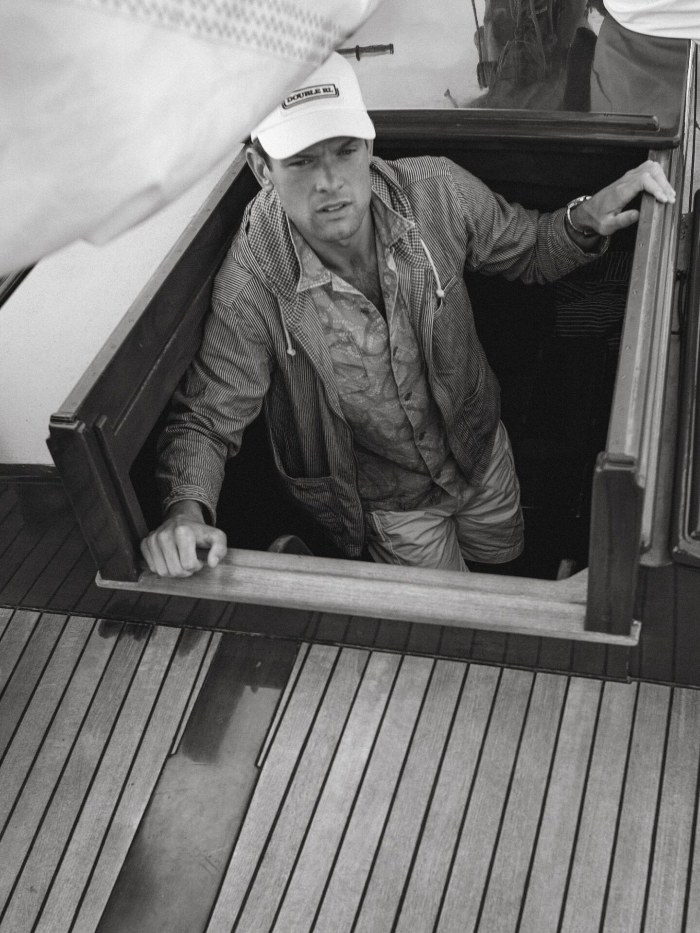 A man emerges from below deck of a boat, wearing a white baseball cap, a hooded shirt, and shorts, with wood decking visible.