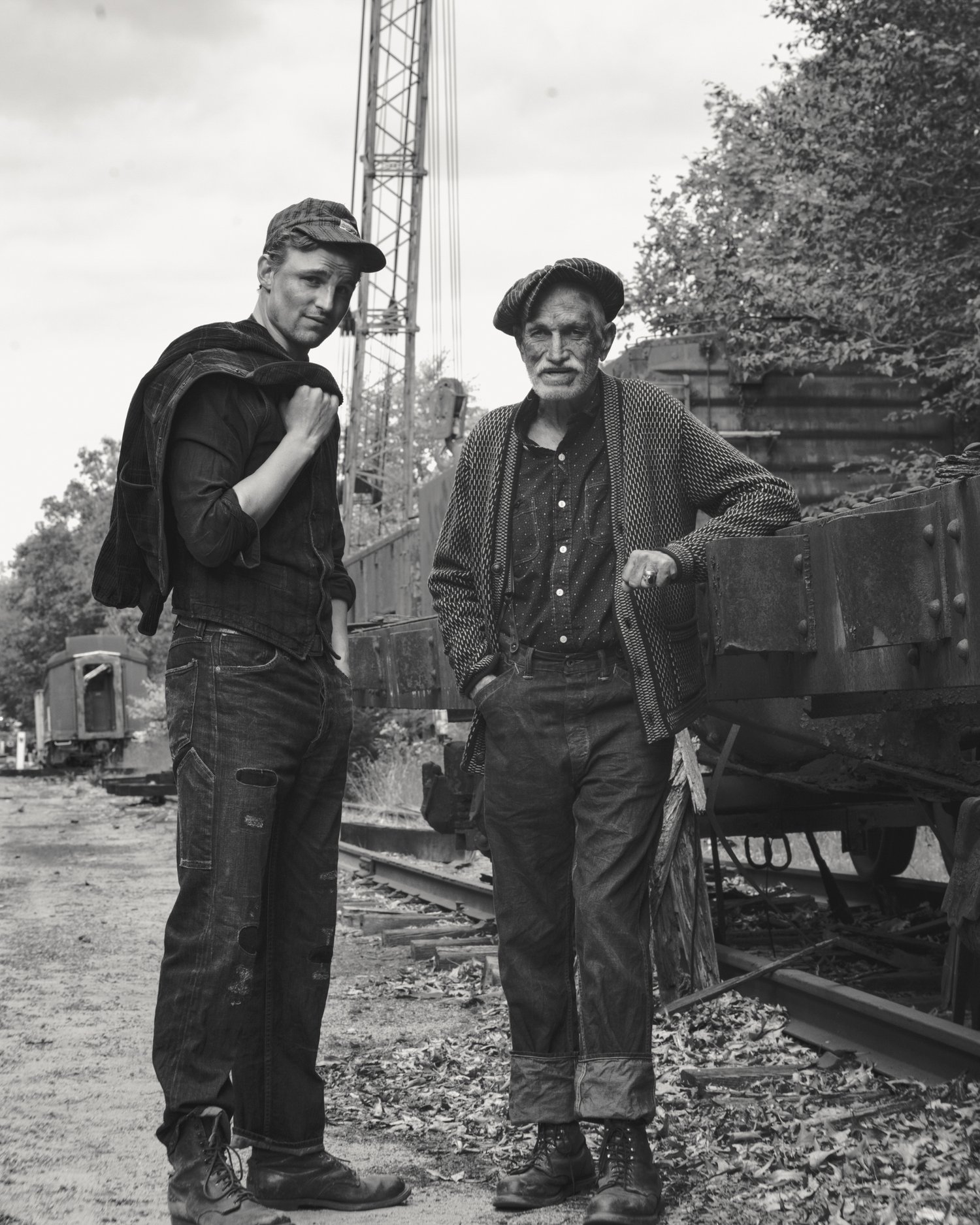 Monochrome portrait of two men in work clothes outdoors with a crane visible in the background, exuding a sense of heritage and labor.