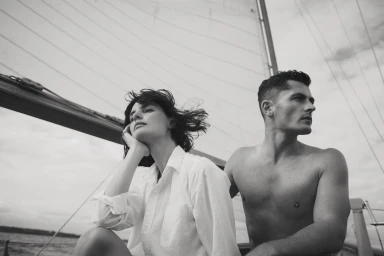 Black and white portrait of a woman in a white shirt and a shirtless man on a sailboat under an overcast sky.