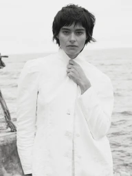 Black and white portrait of a woman with short dark hair wearing a white high-neck jacket by the sea.