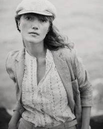 Black and white portrait of a woman wearing a newsboy cap, tailored linen blazer, and patterned ruffled blouse outdoors.