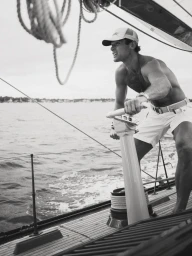 Man wearing a baseball cap and light shorts operating a sailboat winch on open water.