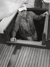Man wearing a cap and jacket emerging from the cabin hatch of a wooden sailboat.
