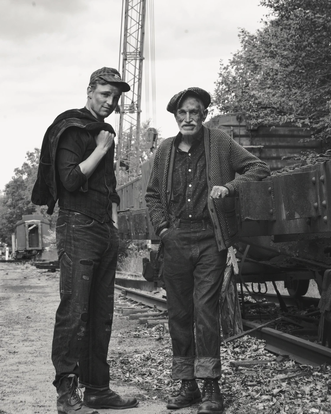 Monochrome portrait of two men standing by a railway line with industrial rail cars and a crane in the background.