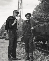 Monochrome portrait of two men standing by a railway line with industrial rail cars and a crane in the background.