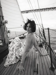 Woman in patterned halter dress sitting on wooden sailboat deck looking back at camera under bridge