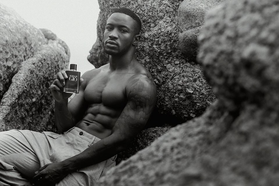 Shirtless man with muscular physique posing with a cologne bottle on rocky beach in black and white, showcasing grooming and fitness.