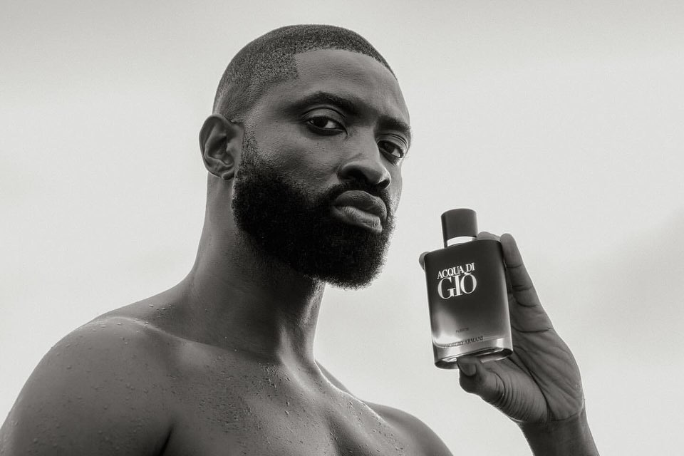 Close-up black and white shot of a Black adult male holding a bottle of Acqua di Gio fragrance, set against a bright background.