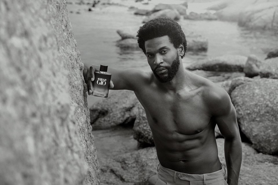 Black and white portrait of a muscular Black male model posing shirtless with a bottle of Acqua di Gio perfume on a rocky beach.