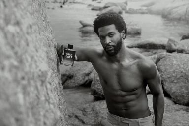 Black and white portrait of a shirtless Black man leaning against a rock on a rocky coast holding a perfume bottle.