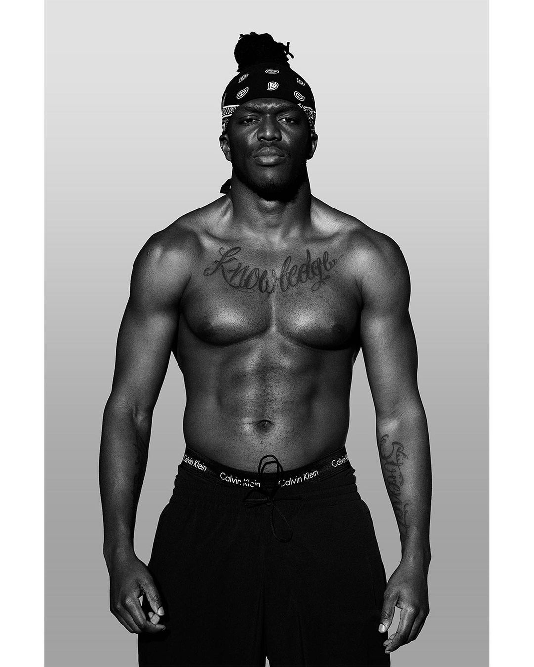 Black and white portrait of a shirtless man with tattoos and a bandana, showcasing fitness and physique against a neutral grey background.