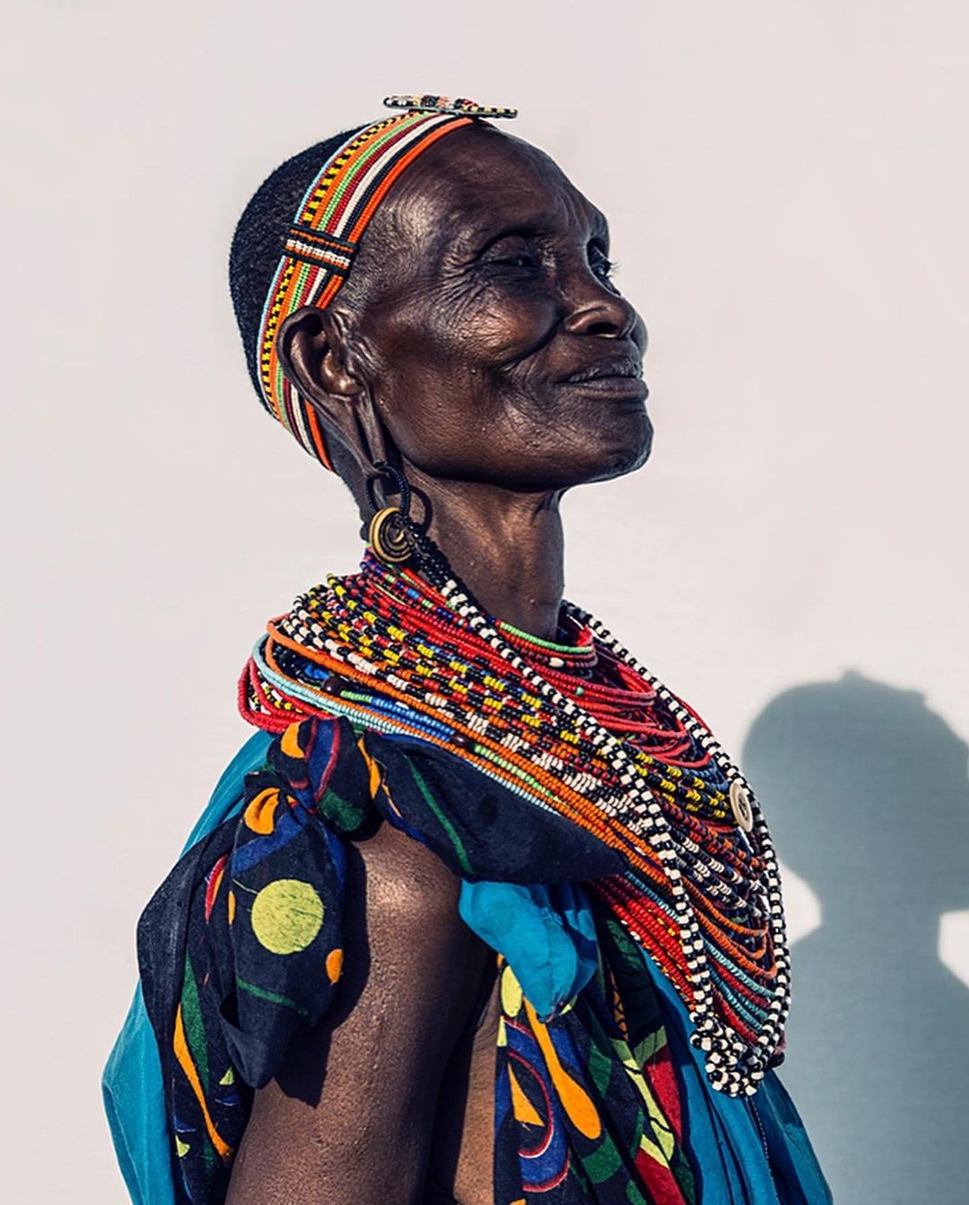 Portrait of an elderly African woman adorned with traditional beaded jewelry and headwear against a bright background, looking upwards.