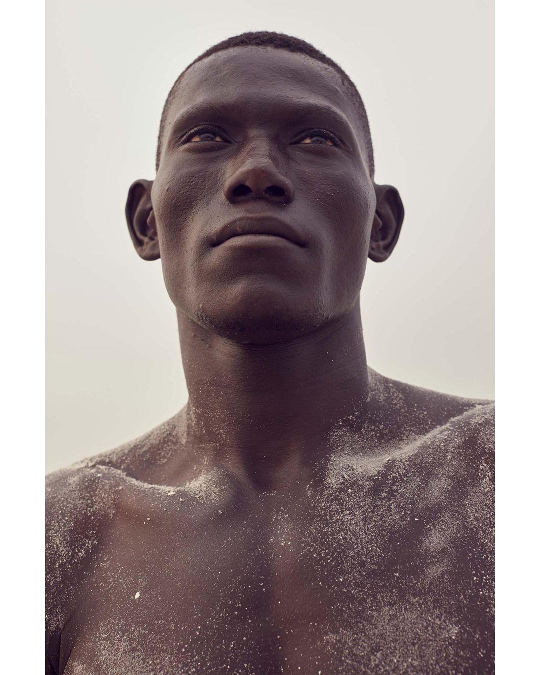Portrait of a dark-skinned adult male looking upwards, covered in white sand on his face and upper body against a light background.