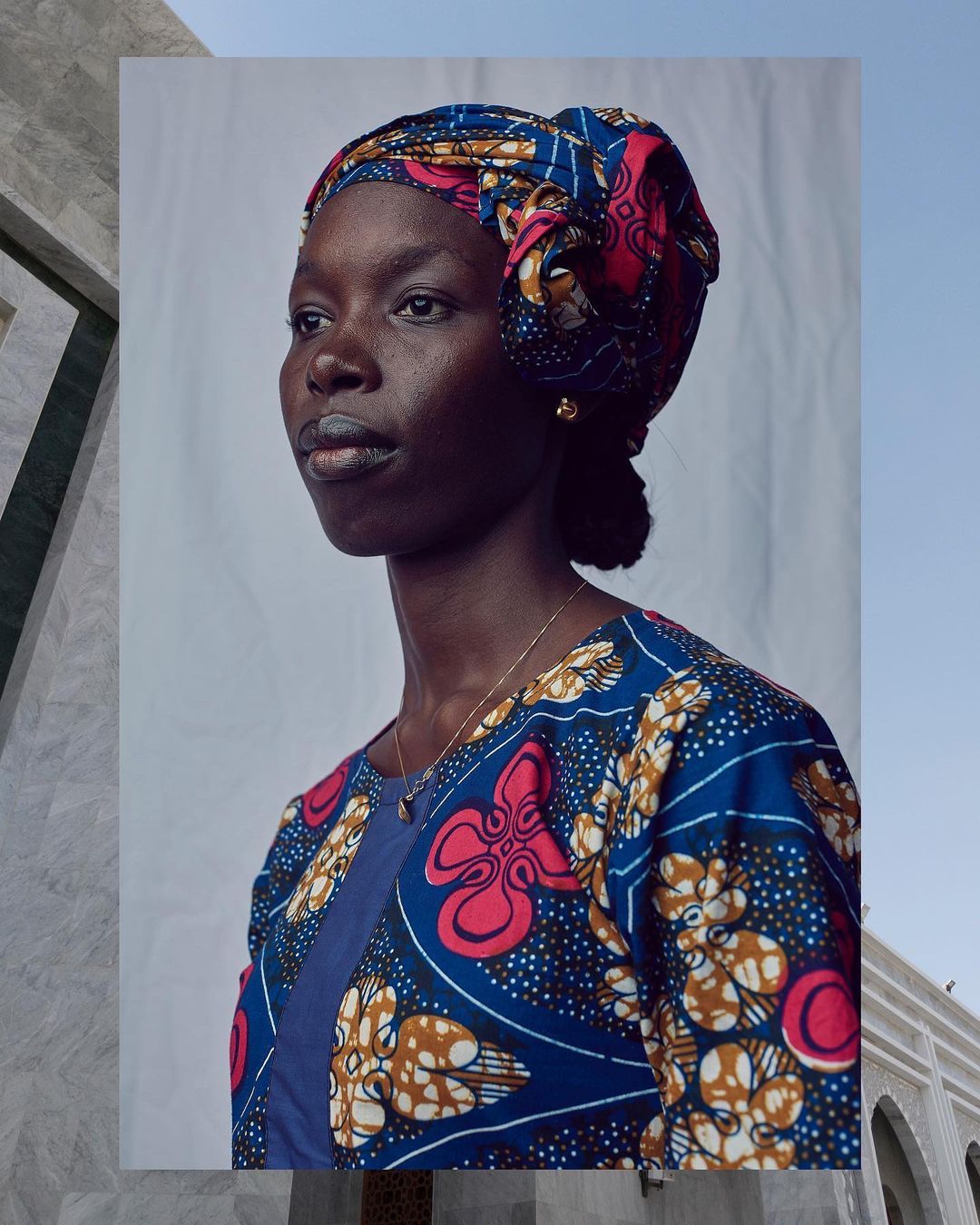 Portrait of a Black woman wearing a colorful patterned headscarf and top against a white backdrop with partial architectural elements.