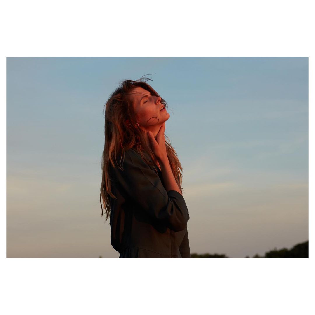 Woman with long hair stands outdoors in natural light with a blue sky background, eyes closed, in a portrait orientation.