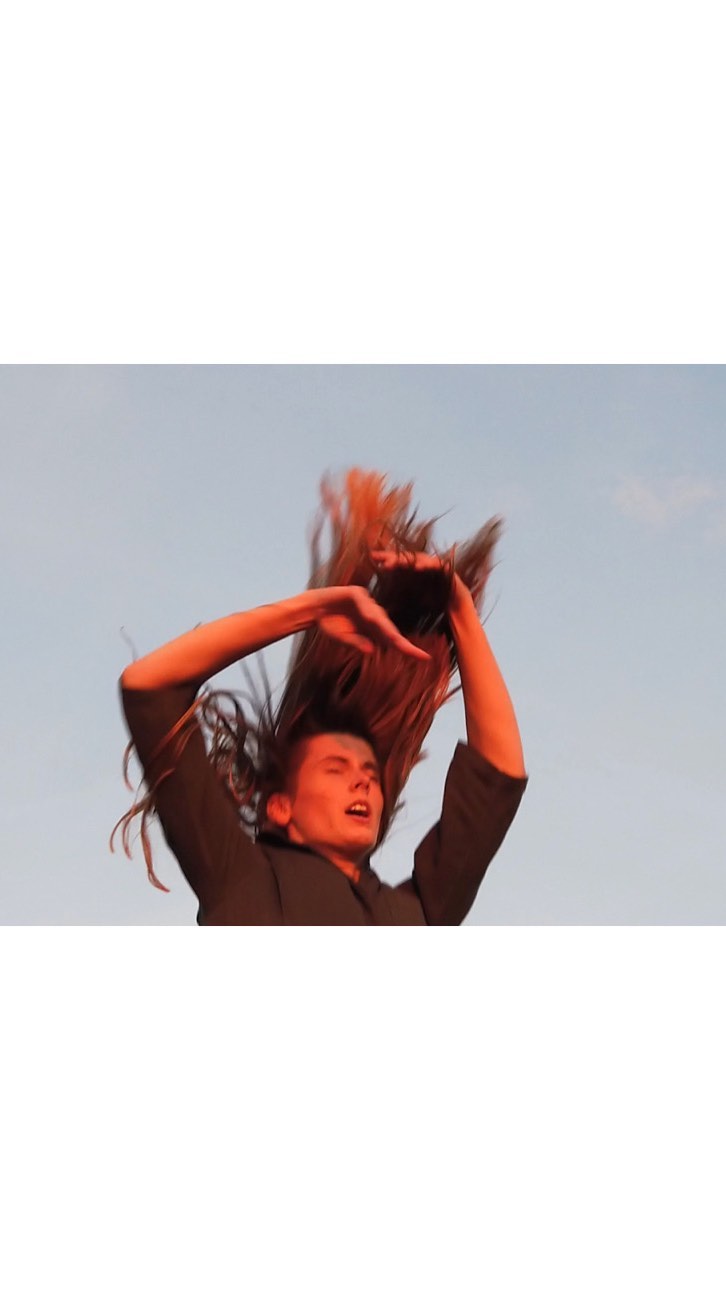 Adult with long brown hair tosses it over their head while posing against a clear sky during daytime with neutral and orange lighting.