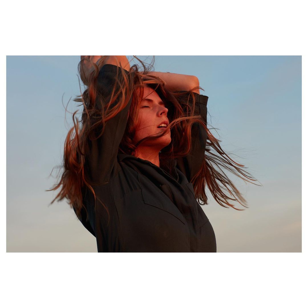 A young woman with long red hair raises her hands to the sky, lit by golden sunlight in an outdoor setting.