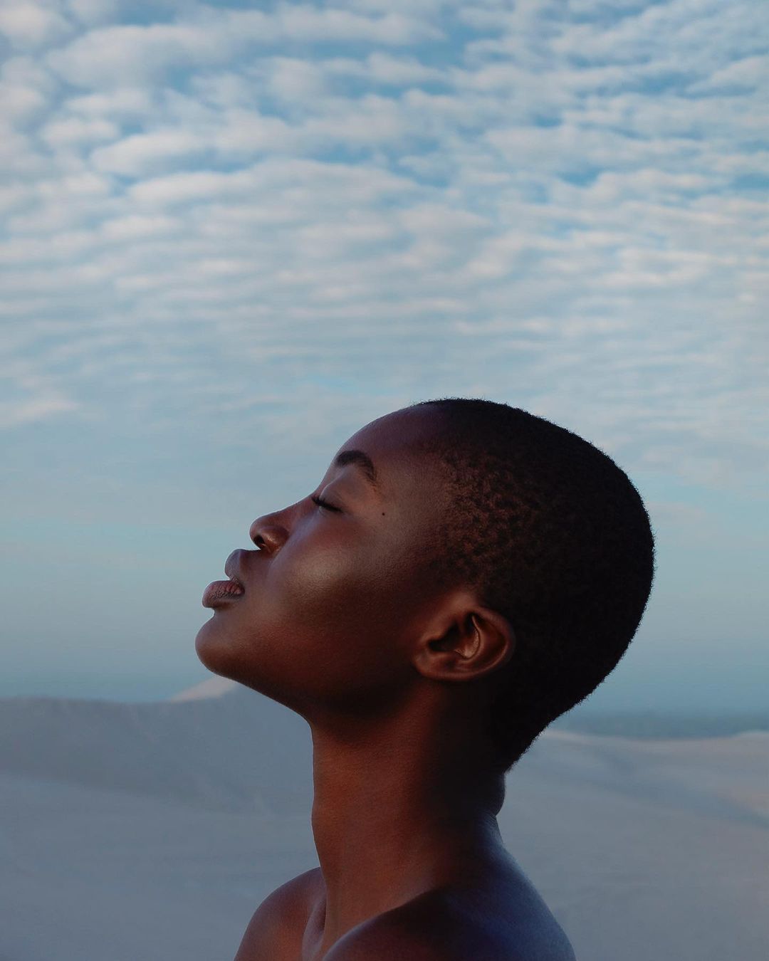 Portrait of an African woman with short hair in profile, eyes closed, outdoors with sky and sand dune background, minimalist beauty.