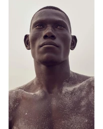Close-up portrait of a Black man with sand dusting his bare skin against a pale sky background