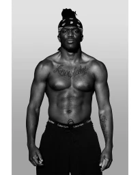Monochrome portrait of KSI standing shirtless wearing a patterned bandana and dark trousers against a plain gradient background.