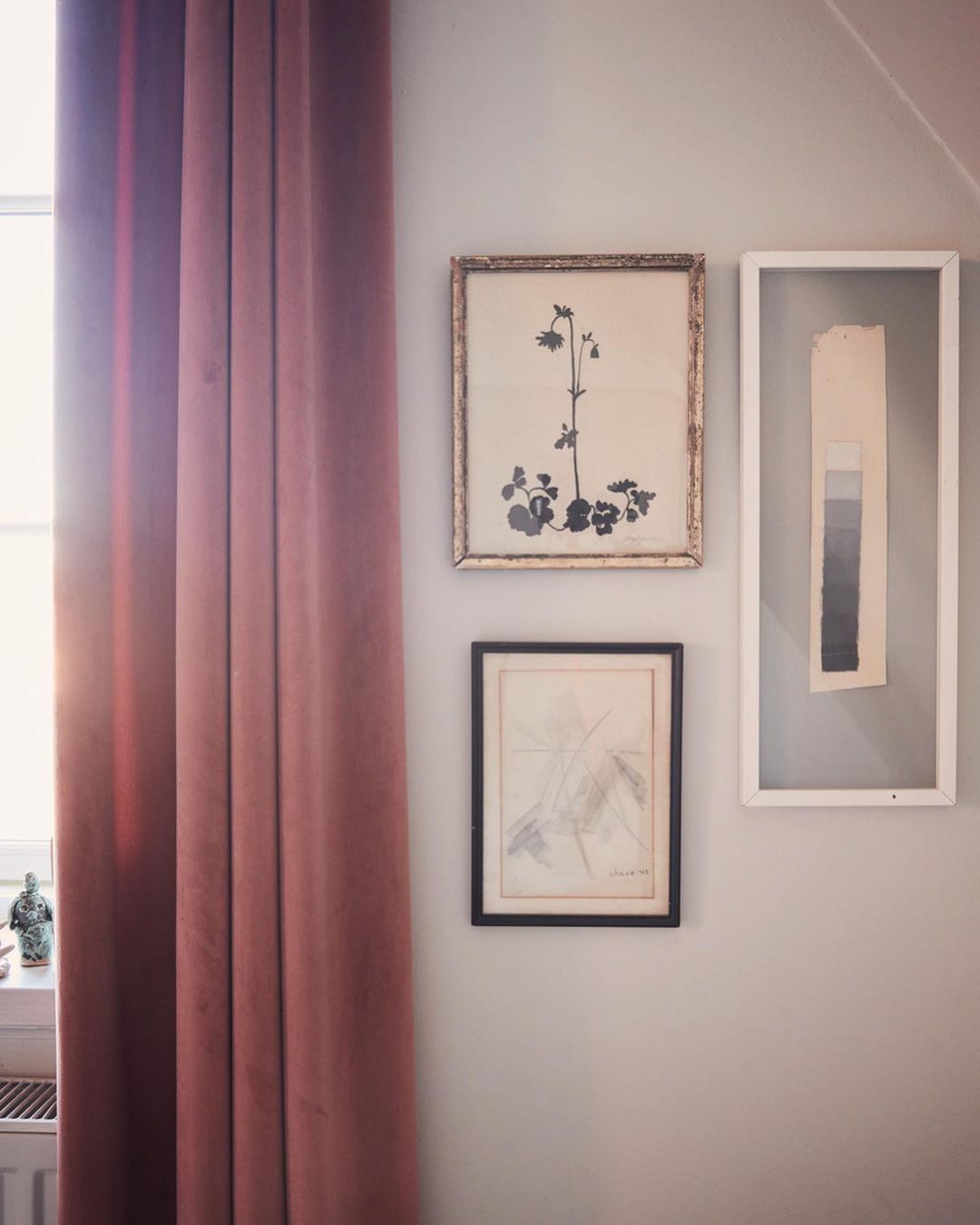 Interior view of a light gray wall decorated with three framed art pieces, with a soft pink curtain on the left side and a window.