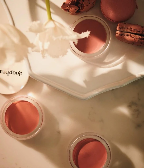 Overhead shot of cosmetic cream in jars, white tulip, and macarons on a white marble surface, highlighting product placement and still life.