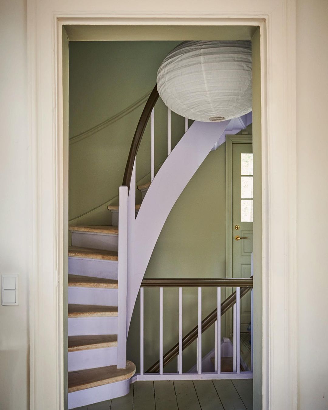 Interior view of a curved staircase with pale lavender accents and a paper lantern, seen through a doorway. Calm, inviting home setting.