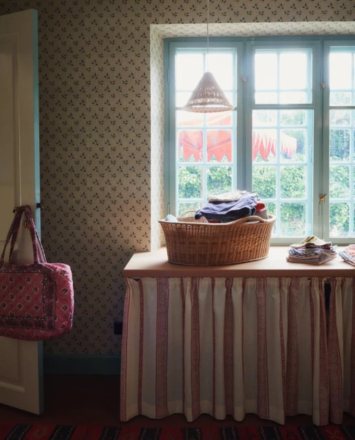 Sunlit laundry room with a basket of clothes on a table with a curtain, a vintage aesthetic, and a view out of the window.