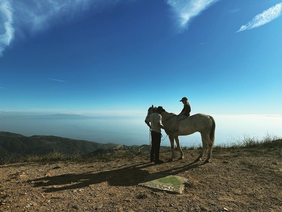 Woman riding a white horse on a hilltop, with an adult male handler, against a background of blue sky, ocean, and distant mountain ranges.