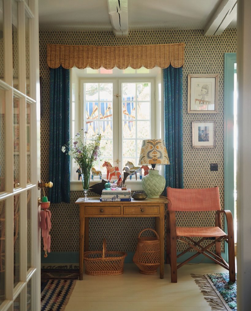 Charming interior with wooden table, flowers, horse figurines, and patterned wallpaper near a window with blue curtains and decor.