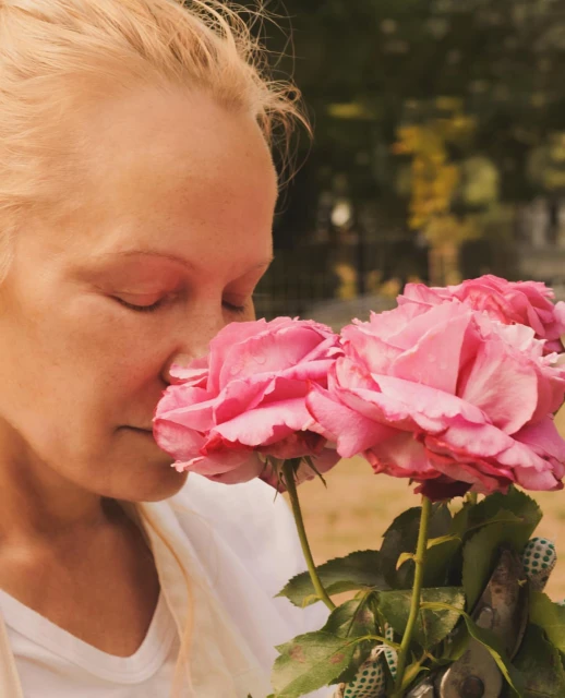 A woman with blonde hair is seen in a close-up, eyes closed, smelling a bouquet of pink roses. She is wearing gardening gloves.