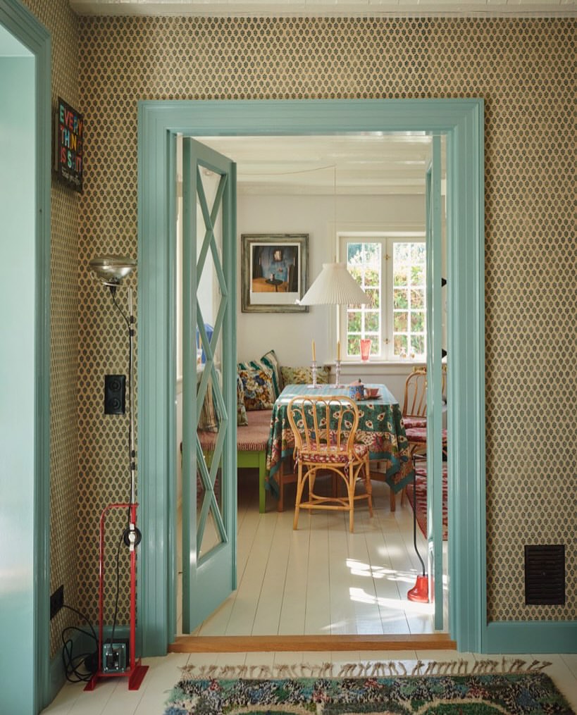 View into a brightly lit dining room from a patterned-wallpapered hallway through a turquoise framed doorway with opened glass paneled doors.