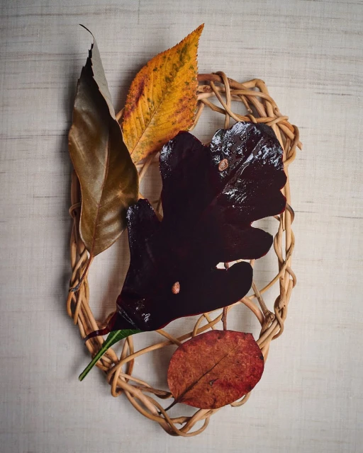 Still life of brown, yellow, and red leaves arranged on a woven, light-brown wreath against a light, wood-grain background.
