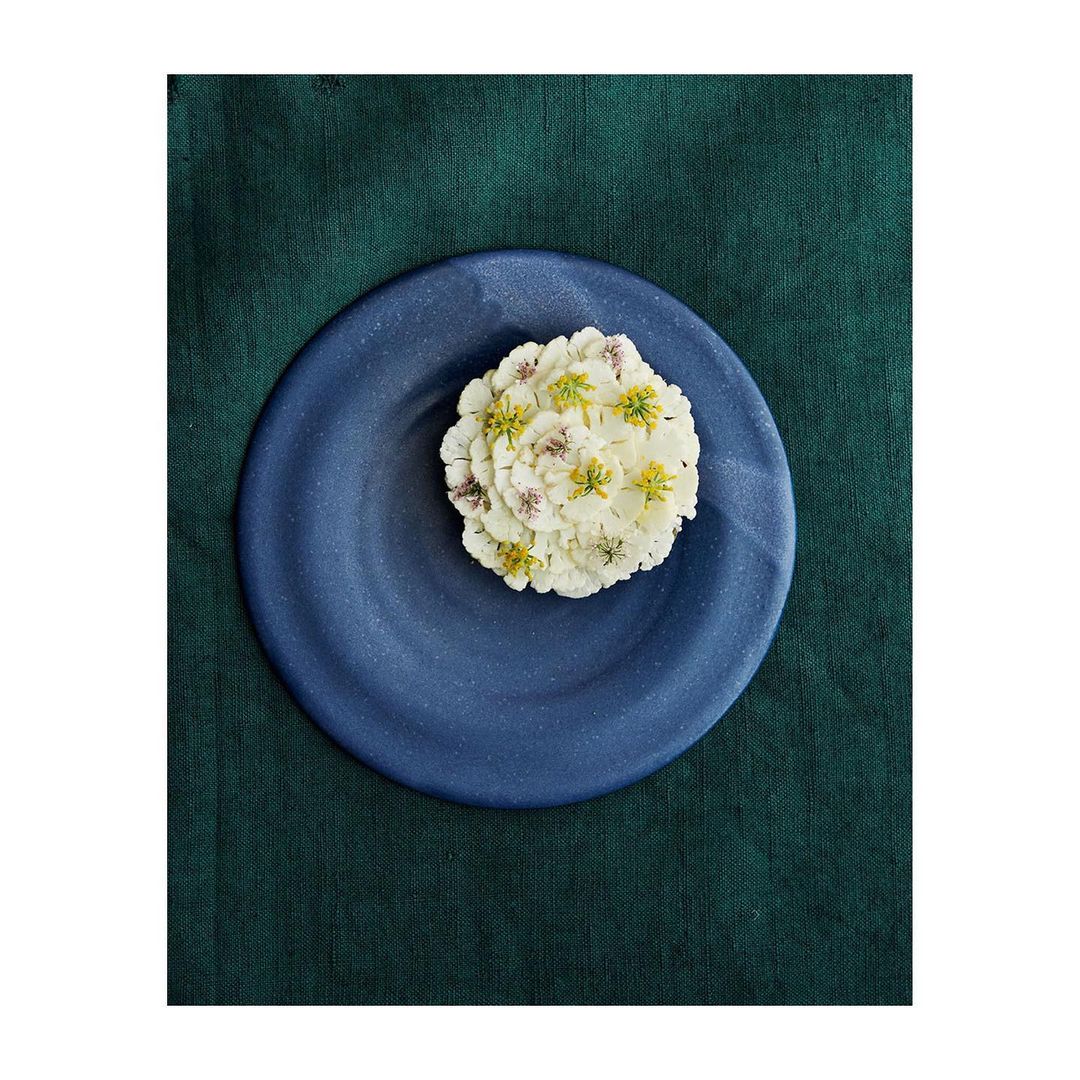 Overhead shot showcases a navy blue plate holding a small white edible flower arrangement, set against a dark green textile backdrop.