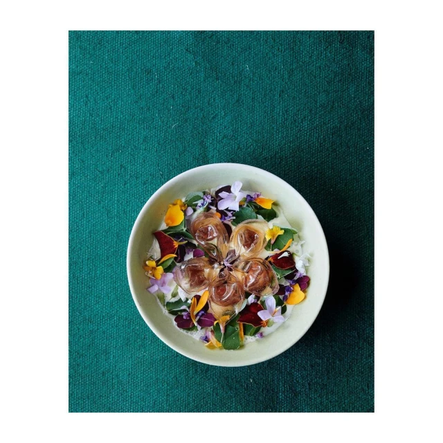 Overhead shot of a white bowl with ice cubes, purple, yellow, and green edible flowers, set against a green fabric backdrop.
