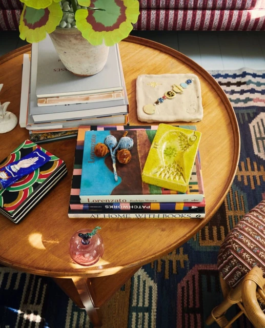 Still life on a round wooden table featuring stacked books, a flowerpot with greenery, and a small pink glass container indoors.
