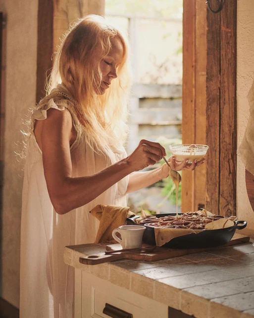Woman in a white dress icing cinnamon rolls in an indoor kitchen near a window, creating a warm and inviting culinary scene.