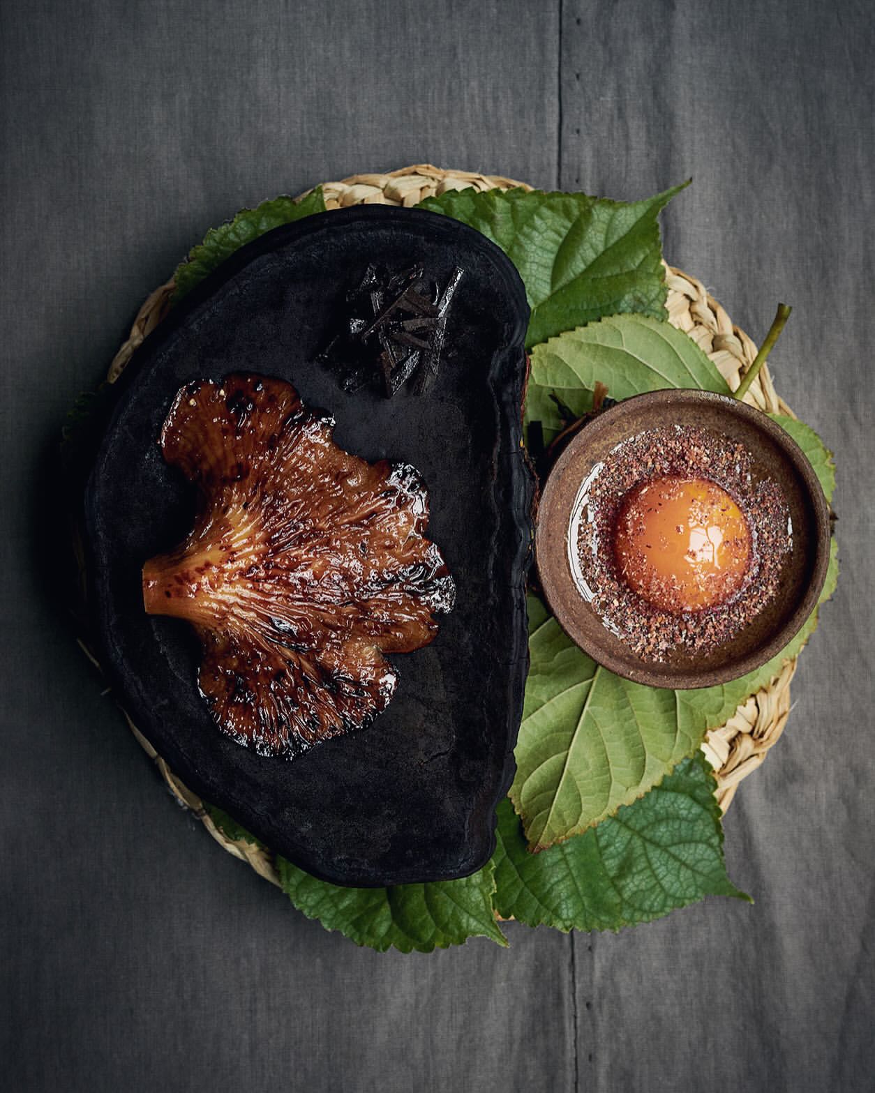 Overhead close-up of a brown cooked mushroom on black pottery beside an orange yolk in a bowl, resting on green leaves and straw.