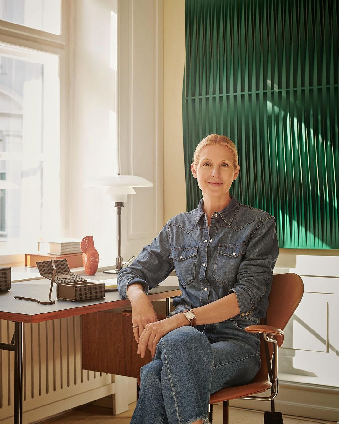 Adult woman poses indoors near desk, wearing a blue denim shirt and jeans. The setting includes a modern lamp and a green art piece.