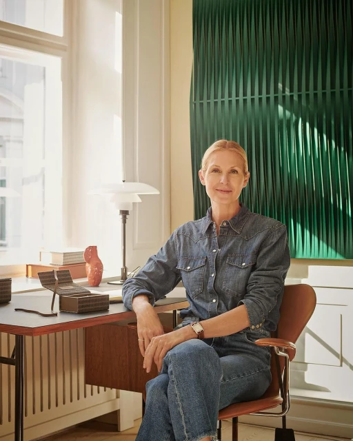 Adult woman poses indoors near desk, wearing a blue denim shirt and jeans. The setting includes a modern lamp and a green art piece.
