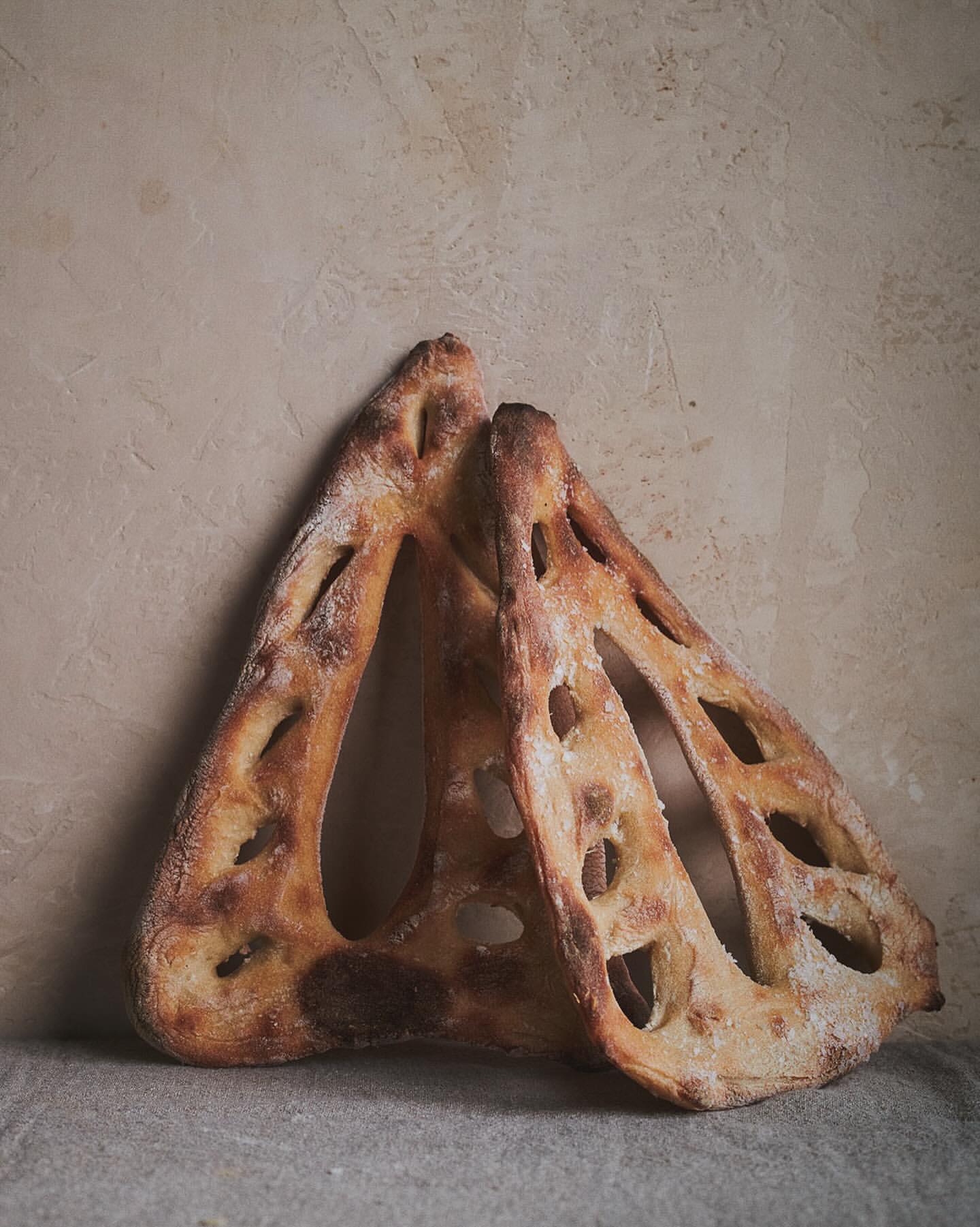 Two loaves of pane bucheron bread with a lattice cut pattern, arranged on a linen cloth against a textured wall. Close-up shot.