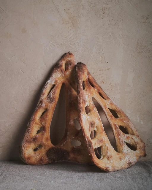 Two loaves of pane bucheron bread with a lattice cut pattern, arranged on a linen cloth against a textured wall. Close-up shot.