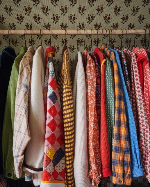 A collection of colorful vintage clothing items on a metal rod with a floral wallpaper backdrop in an indoor setting.