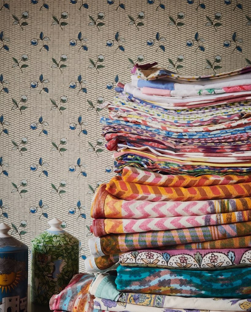 Stack of colorful folded textiles and fabric with floral wallpaper behind, along with two ceramic jars on a dark surface.