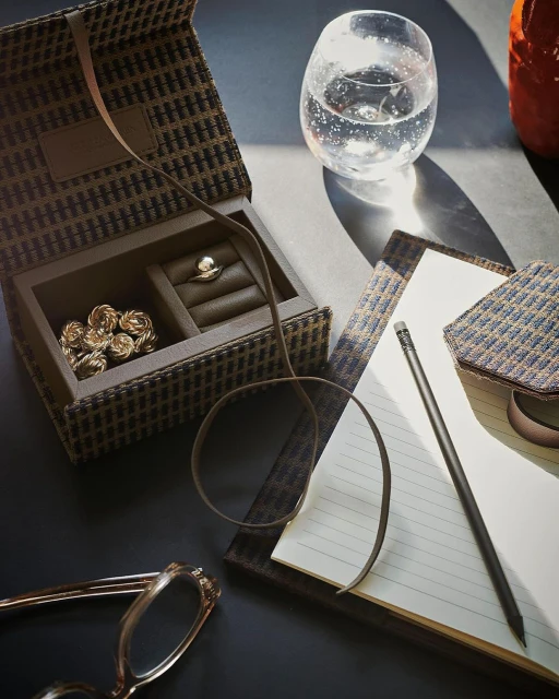 Still life composition featuring a jewelry box with gold rings, a glass of sparkling water, eyeglasses, a notebook, and a pencil on a dark surface.