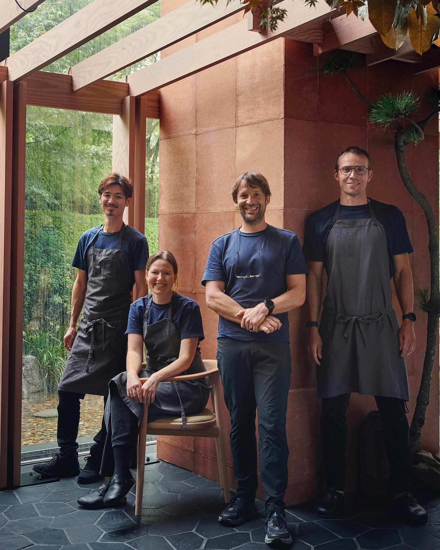 Group of four restaurant employees in aprons posing indoors next to a glass wall overlooking greenery and a stone wall.
