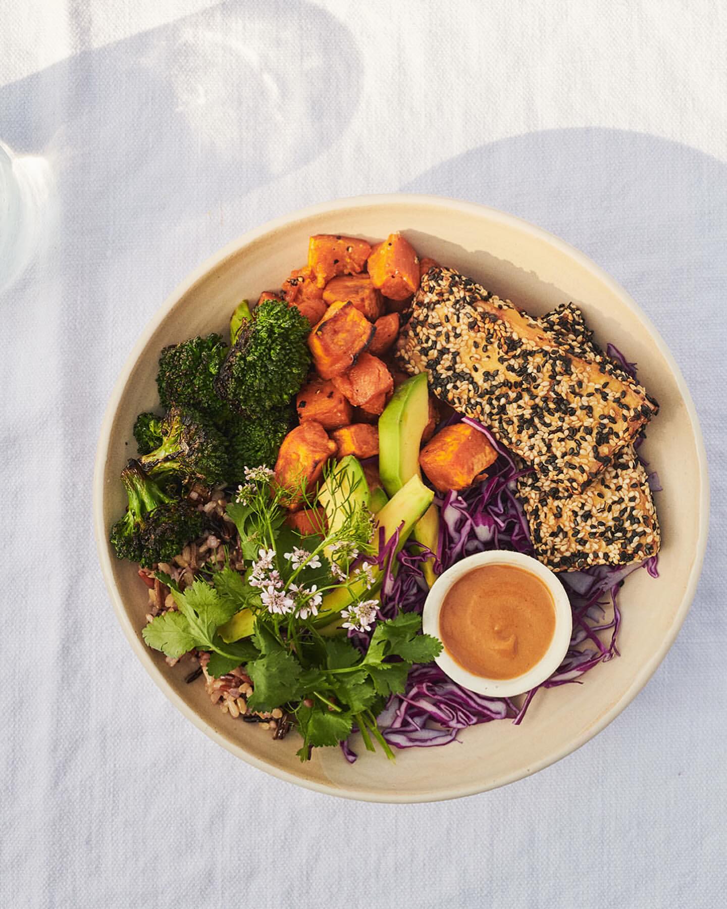 Overhead shot of a vibrant grain bowl with broccoli, sweet potato, avocado, red cabbage, cilantro, and sesame-crusted tofu on a white tablecloth.