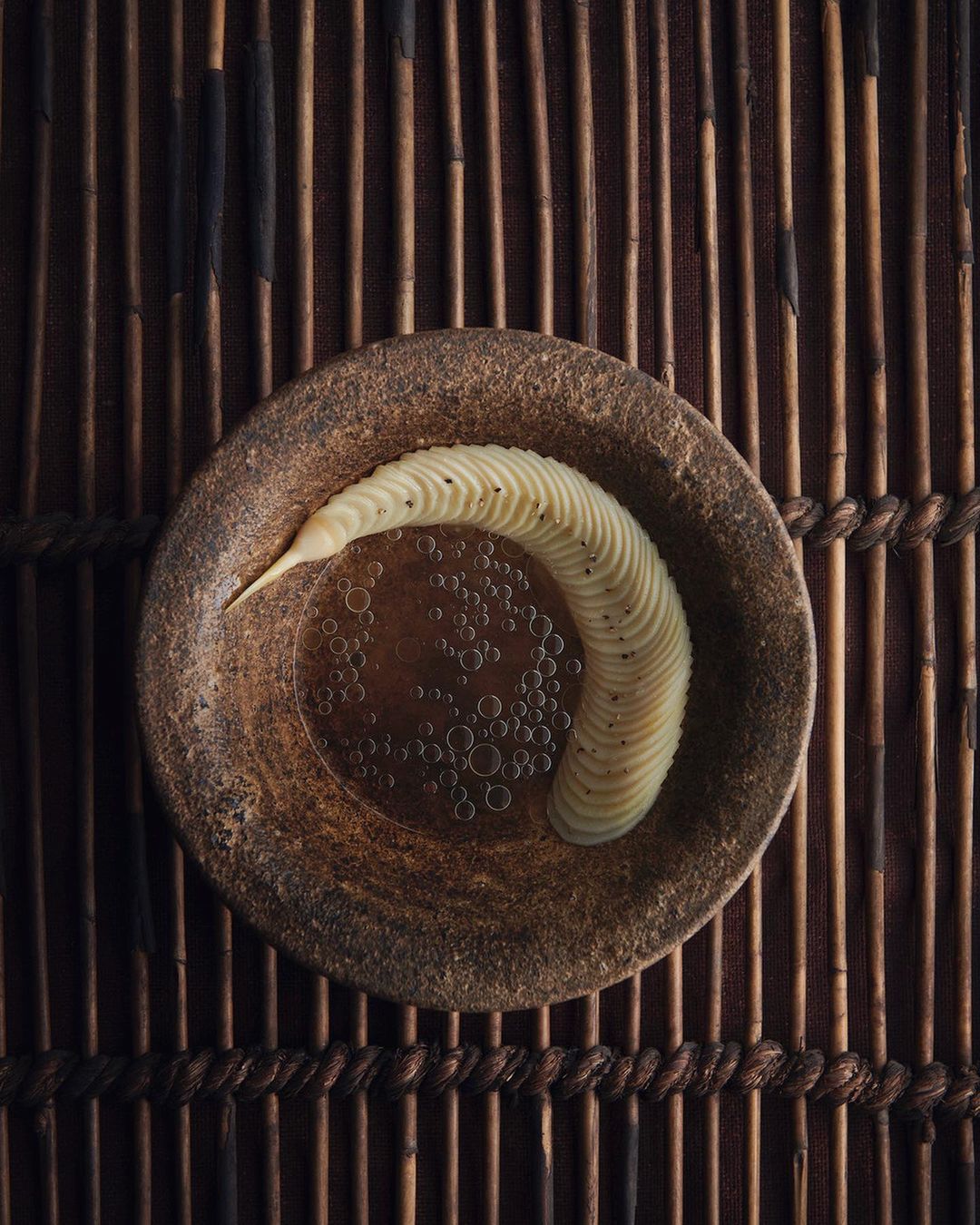 Overhead shot of spiral sliced potato dish with clear broth on a speckled brown plate, set against a rustic bamboo background.