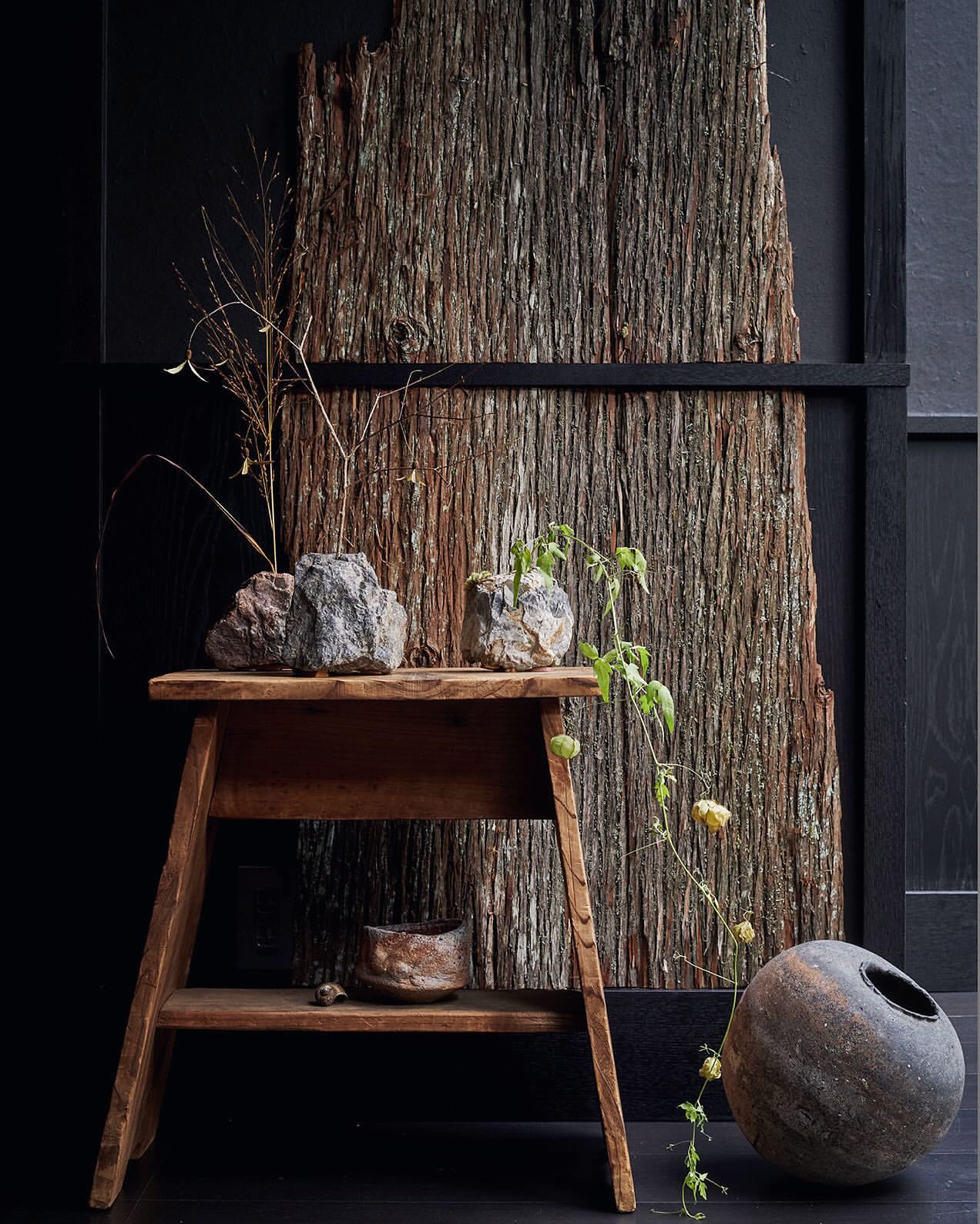 Still life composition featuring a rustic wooden stool, stone planters with greenery, and a textured wooden backdrop against a dark wall.